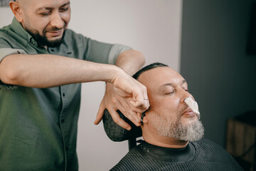 Male barber trimming male client's ear hair with nasal wax absorbent