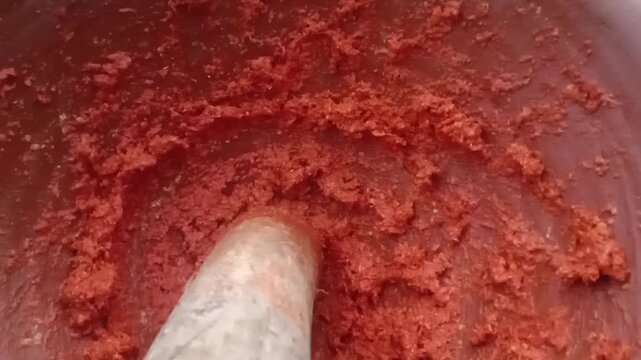 Closeup of red chili paste being ground on a traditional stone mortar and pestle showing rich texture vibrant color and natural spices blending into a smooth aromatic mixture
