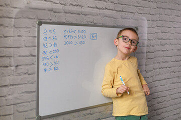 A confident six-year-old boy in glasses, wearing a yellow shirt and green pants, stands on the right side of a whiteboard and points to corrected math examples. Concept of early learning confidence.