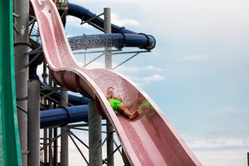 A person in bright green swim trunks gleefully journeys down a winding pink water slide at a park, water splashing all around. A fun, sunny scene