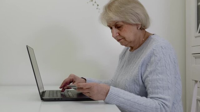 Senior woman in gray cable-knit sweater working on laptop from side view. Focused typing showing productivity, digital work and technology proficiency in bright home office setting.