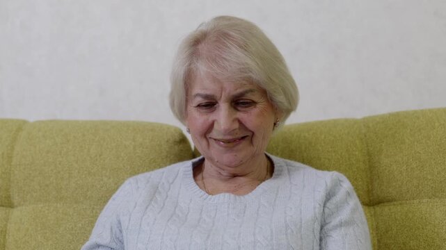 Elderly woman in gray knit sweater using smartphone with genuine happy smile on yellow sofa. Positive emotion showing enjoyable online content, digital communication and technology satisfaction.