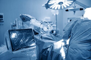 In a sterile Operating Room, a surgeon in scrubs uses a robotic system. The robotic arms hover over the patient, while the monitor displays vital information during surgery