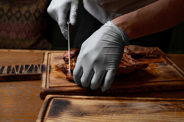 Chef cutting grilled pork ribs on wooden board, closeup of hands in gloves slicing juicy roasted meat with knife, barbecue preparation process in restaurant kitchen
