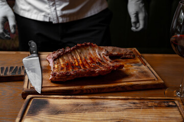 Grilled pork ribs on wooden board with chef in background, sharp knife and glass of red wine, rustic barbecue serving in restaurant kitchen