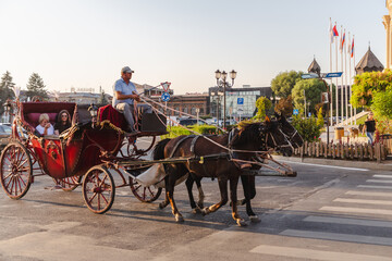 Gyumri, Armenia - 24 August 2025: View of a horse-drawn carriage with passengers crossing a sun-drenched street, framed by buildings and flags against a clear sky.
