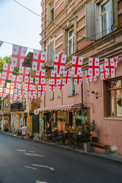 Tbilisi, Georgia - 19 August 2025: View of the vibrant street adorned with Georgian flags, casting shadows on the old pink buildings and inviting cafes below.