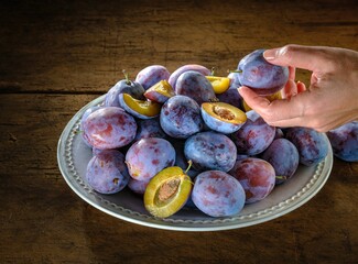 A hand picks a ripe plum from a luscious heap on a charming plate. Some plums are already sliced, revealing their golden flesh, on a weathered wooden surface © Damian