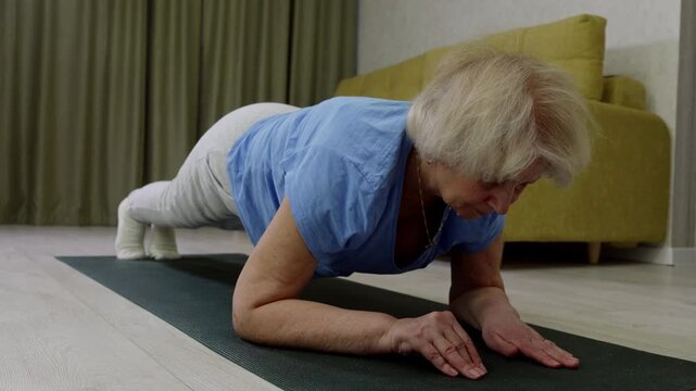 Senior woman in blue t-shirt holding forearm plank position on green yoga mat. Challenging core exercise showing determination, proper form and fitness dedication at home.