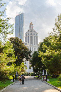 Batumi, Georgia - 21 August 2025: View of the sky-piercing skyscraper and the Radisson Blu Hotel Batumi, framed by lush green trees, as a couple strolls along the sun-dappled path.