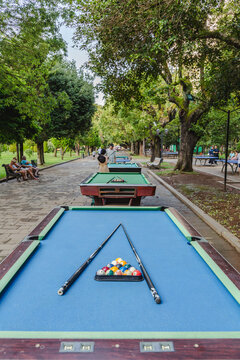 Batumi, Georgia - 21 August 2025: View of vibrant billiard tables lined along a stone pathway, framed by lush trees and benches in a tranquil park setting.