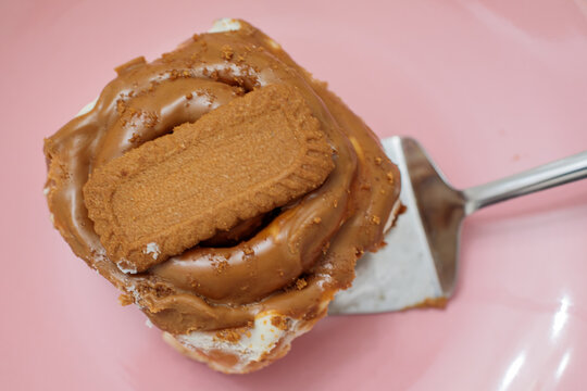 Freshly baked cinnamon roll topped with creamy biscoff spread and a lotus cookie, being served on a metal spatula on a glossy pink plate