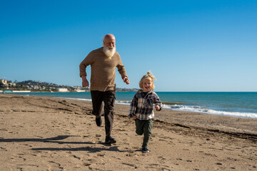 Healthy active senior man running along the beach with his young grandson on a sunny day, enjoying family time, outdoor activity
