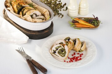 Sliced turkey roll, stuffed with verdant herbs, lies on a white plate garnished with red berries. A baking dish and wooden platter of carrots add to the rustic, autumnal scene