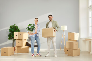 Full length portrait of happy young couple standing with a lot of cardboard boxes in a new apartment and looking at camera celebrating moving day. Relocating, real estate, mortgage concept.
