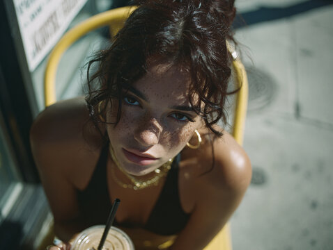 a person sitting outdoors, holding a beverage in a clear cup with a straw. They are wearing a black tank top and layered gold necklaces. The background features a stone building with a window