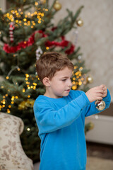 Cute little boy in a blue sweater decorating a christmas tree with a gold ornament in a cozy home interior, preparing for winter holidays and new year celebration