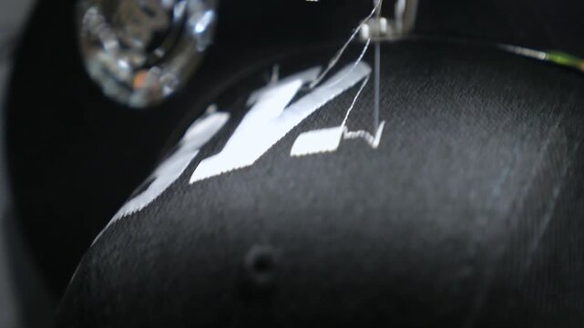 Detailed macro view of an automated embroidery machine stitching white thread lettering onto a black fabric baseball cap, showcasing the modern textile industry and custom apparel manufacturing
