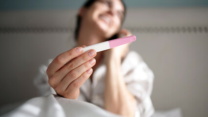 Woman holding pregnancy test with positive result, smiling while sitting on bed, surrounded by soft bedding, conveying joy and anticipation of new beginnings
