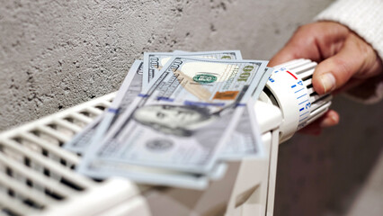Hand adjusting thermostat with dollar bills placed on top, symbolizing rising energy costs and...