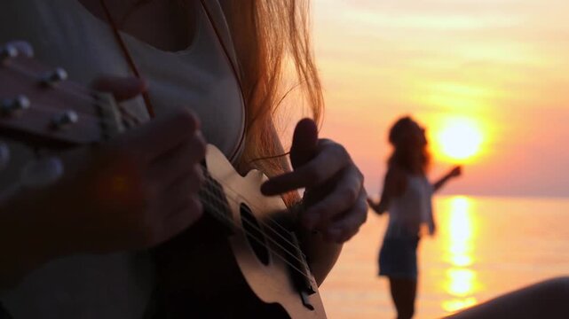 Close up of a young woman playing a ukulele on the beach at sunset while her friend dances joyfully in the background, their silhouettes creating a beautiful, warm and happy summer scene