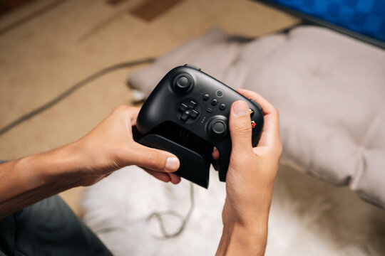 Close-up of unrecognizable person holding modern black wireless game controller in focused home gaming session, hands on buttons and joysticks, enjoying casual digital entertainment, competitive play.
