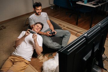 Gay couple spending quality time together, one man lying on floor concentrating on video game, other resting in bean bag chair watching large TV screen.