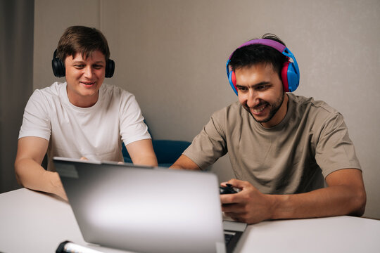 Portrait of two cheerful young men gathered in living room, laughing and focused on laptop and controllers while gaming together, showcasing friendship, teamwork and casual digital leisure.