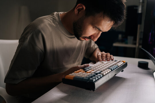 Focused man inspecting custom mechanical keyboard up close, checking keys for dirt and wear, demonstrating careful maintenance, precision and passion for electronics and pc hobbyists.