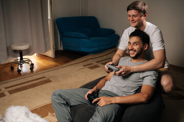 Gay diverse male couple sharing cozy evening gaming at home, one seated with wireless controller while partner leans over to join in, smiling and enjoying playful bonding moment at home.