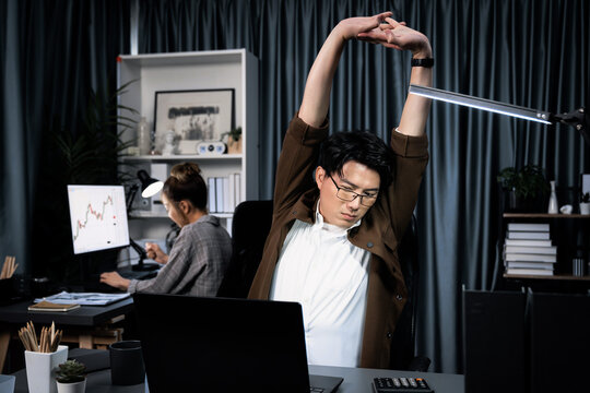 Sales manager working on desk with stretching body with arm ache at over late night time at modern home office with secretary sitting back investing dynamic stock market on project database. Infobahn. - Powered by Adobe