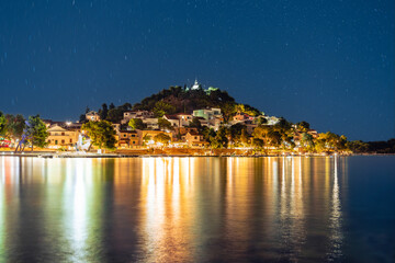 Nighttime panorama of the town of Tribunj in Croatia, featuring a seafront, illuminated promenade, and houses on a hill. The Earth's rotation is visible in the sky with the North Star.