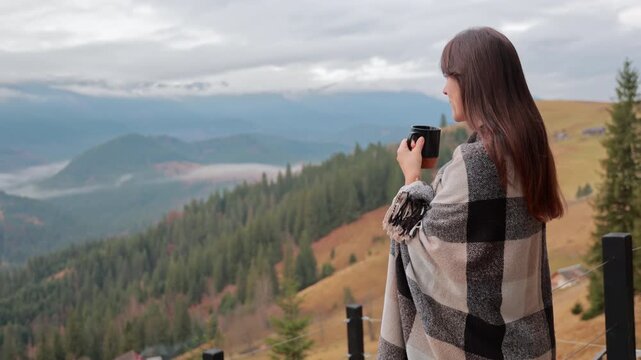 Young woman wrapped in a warm plaid blanket drinking hot tea on the balcony, enjoying the beautiful view of the mountain valley in the autumn