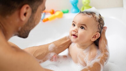 Father gently bathing his baby in a foamy tub as the child smiles up with curiosity and comfort during a warm, playful wash time, everyday family routine, bright mood