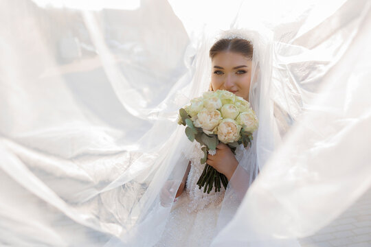 Beautiful bride holding bouquet under veil at outdoor wedding ceremony in soft light