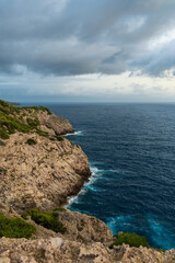 Dramatic view of waves crashing against rocky shoreline under gray storm clouds near Far de Capdepera, Mallorca. Concept of nature power, seascape, rugged coast.