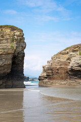 A natural rock arch framed by two massive layered cliffs at las Catedrales Beach, Spain, the wet sand reflects the bright sky and the ocean waves visible through the natural gateway