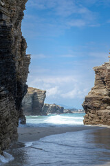 Massive layered rock cliffs frame an ocean view at As Catedrais Beach, Spain. Waves roll onto the wet sand, capturing a majestic geological wonder and famous coastal landmark