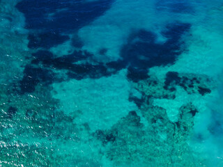 Aerial drone view of transparent sea water at summer. Underwater reefs, sand, rocks and sea grass. Sea waves and sun reflection on water surface. 