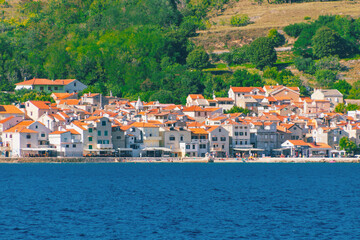 Obraz premium Picturesque coastal village with orange roofs and lush green hills by the sea. Baška, Krk island, Croatia
