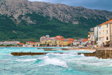 Coastal town with colorful buildings and rocky mountains overlooking turquoise sea waves. Baška, Krk island, Croatia
