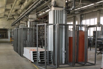 Interior of an industrial warehouse or factory floor with rows of prefabricated metal framing systems and bundled white construction materials near bright windows