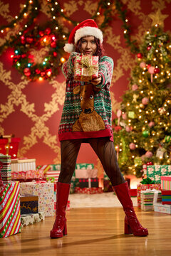 Woman in Santa hat and festive sweater holding wrapped Christmas gift near decorated tree