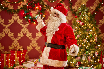 Santa Claus waving in front of decorated Christmas trees with presents and festive holiday lights