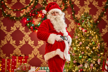Santa Claus holding a smartphone in front of decorated Christmas tree with twinkling lights