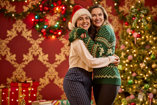 Two women embrace in festive holiday sweaters and Santa hat in front of decorated Christmas tree