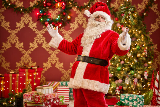 Santa Claus in traditional red suit with white beard standing by decorated Christmas tree with gifts