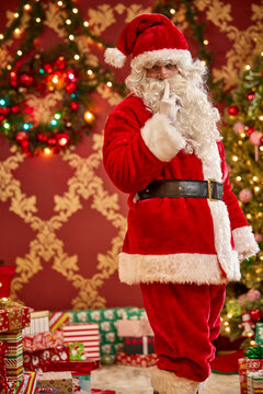 Santa Claus making shush gesture with finger to lips in front of decorated Christmas tree