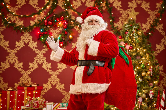 Santa Claus waves while carrying his gift sack in front of decorated Christmas trees