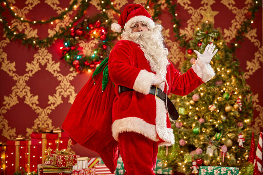 Santa Claus waving with gift sack beside decorated Christmas tree and wrapped presents